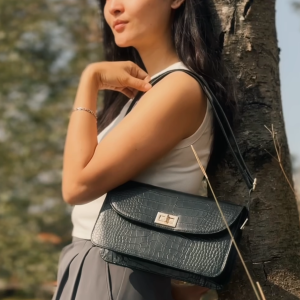 A woman leaning against a tree wearing the Elira Black croc-embossed leather shoulder bag by The Karigar Nepal. The bag features a structured flap design and a gold-tone twist lock closure.