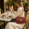 Handcrafted Elira Red genuine leather shoulder bag with a croc-embossed texture and gold hardware, worn by a woman enjoying coffee. The minimalist bag rests elegantly while she holds a latte. Heritage luxury aesthetic from The Karigar Nepal.
