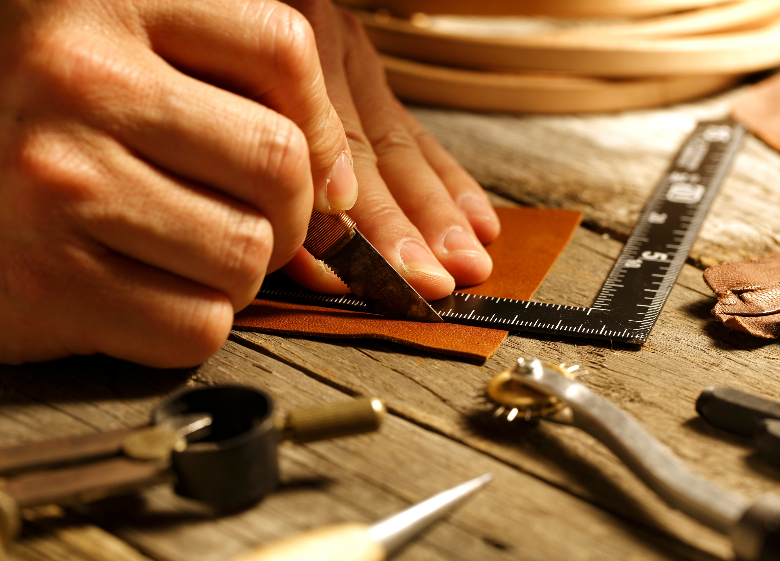 Close-up of a Karigar artisan's hands using a precision knife and a square ruler to hand-cut a piece of premium tan leather on a rustic wooden workbench. Traditional leatherworking tools are scattered in the foreground.