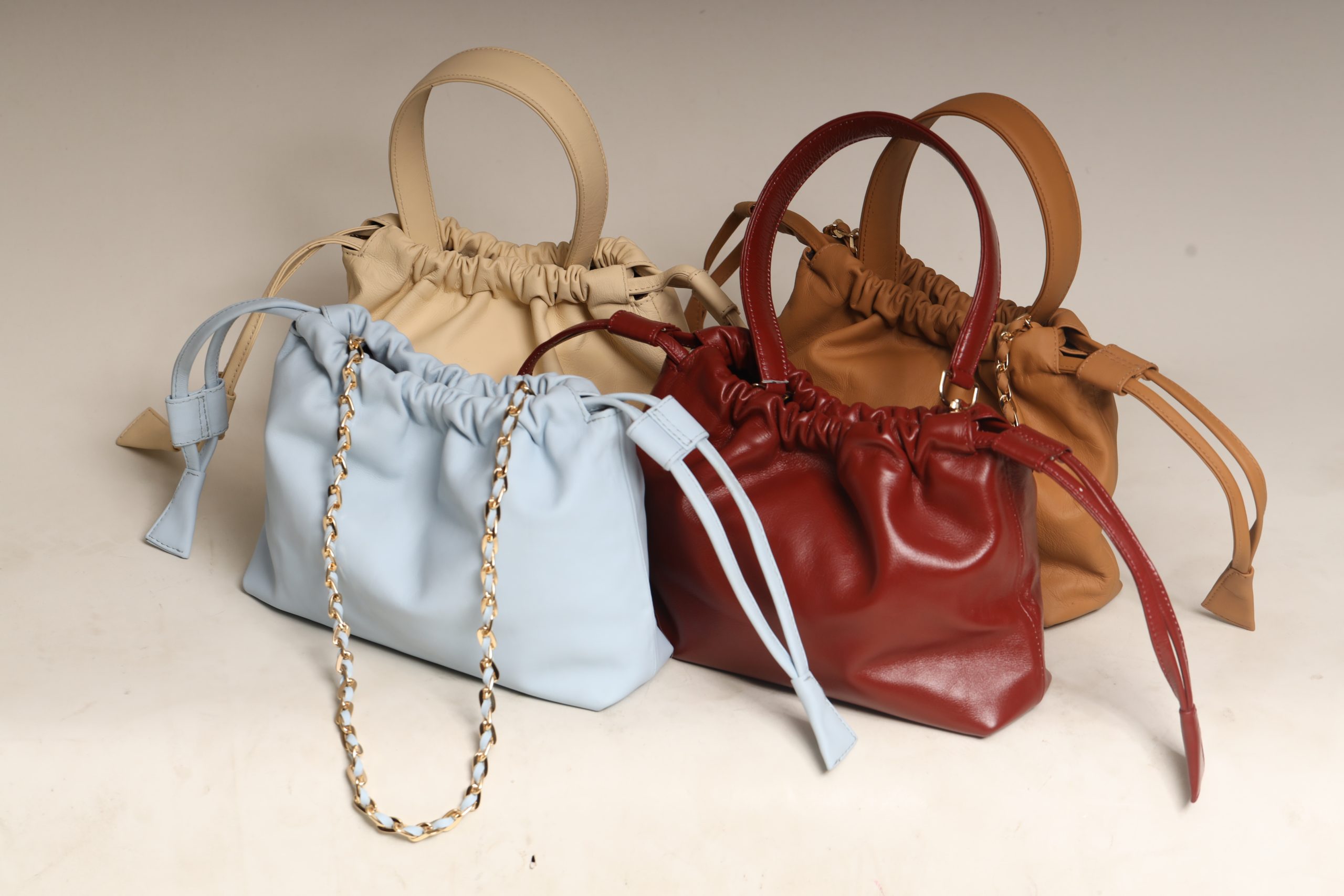 A collection of four handcrafted Thaili leather bags by The Karigar Nepal in Sky Blue, Ruby Red, Camel Tan, and Sand Beige. The bags feature a gathered drawstring design, top handles, and gold-tone chain straps against a neutral background.